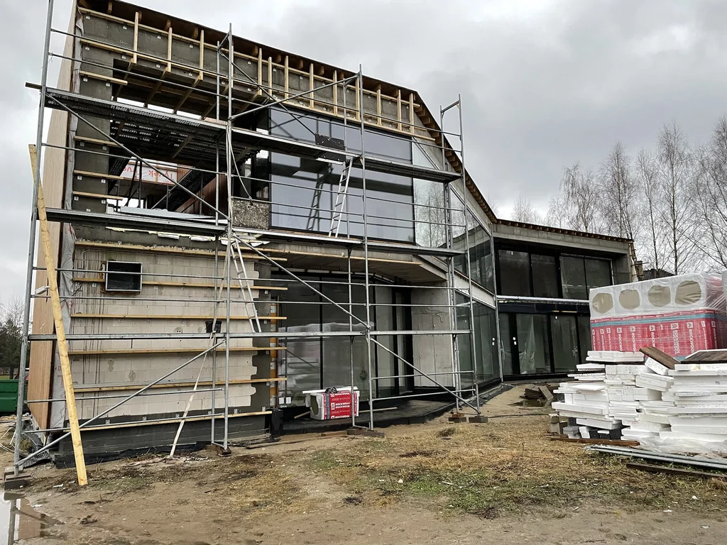 Fasada z dużymi przeszkleniami (w tym narożnymi) oraz rusztowaniem przy montażu elewacji i stolarki.
