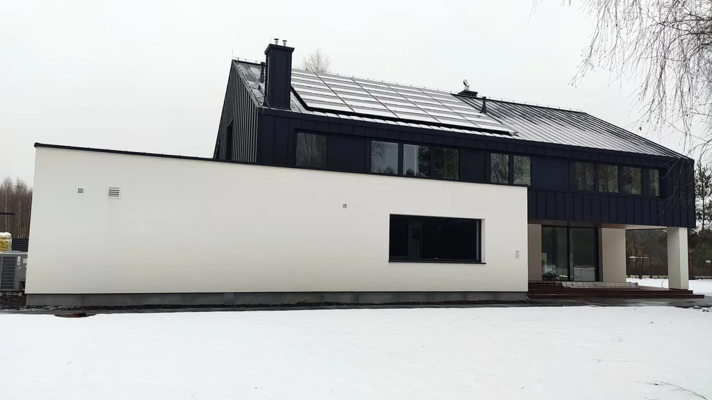 Boczna elewacja nowoczesnego domu zimą, biała bryła i czarny dach z panelami fotowoltaicznymi