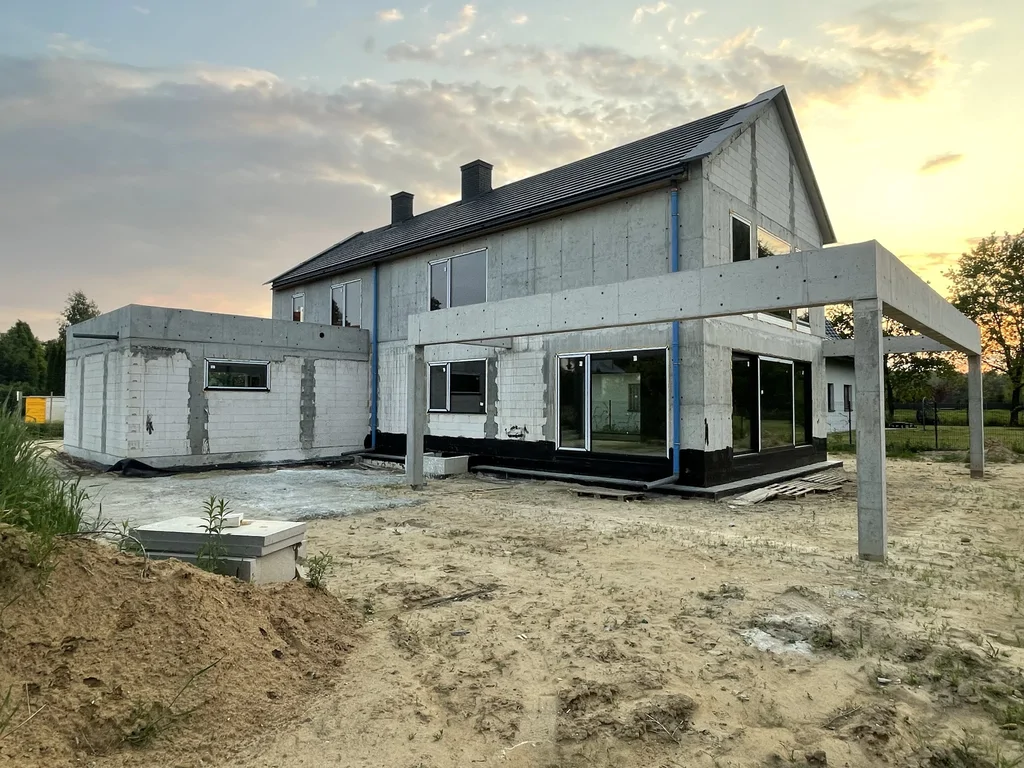Widok domu jednorodzinnego w stanie surowym z żelbetową konstrukcją tarasu i dużymi przeszkleniami o zachodzie słońca