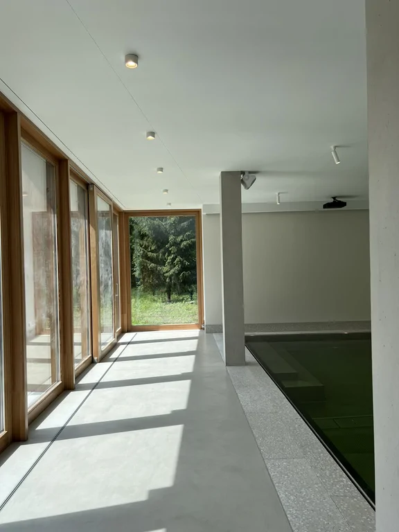 Strefa przybasenowa z wysokimi przeszkleniami, betonowymi słupami i widokiem na zieleń