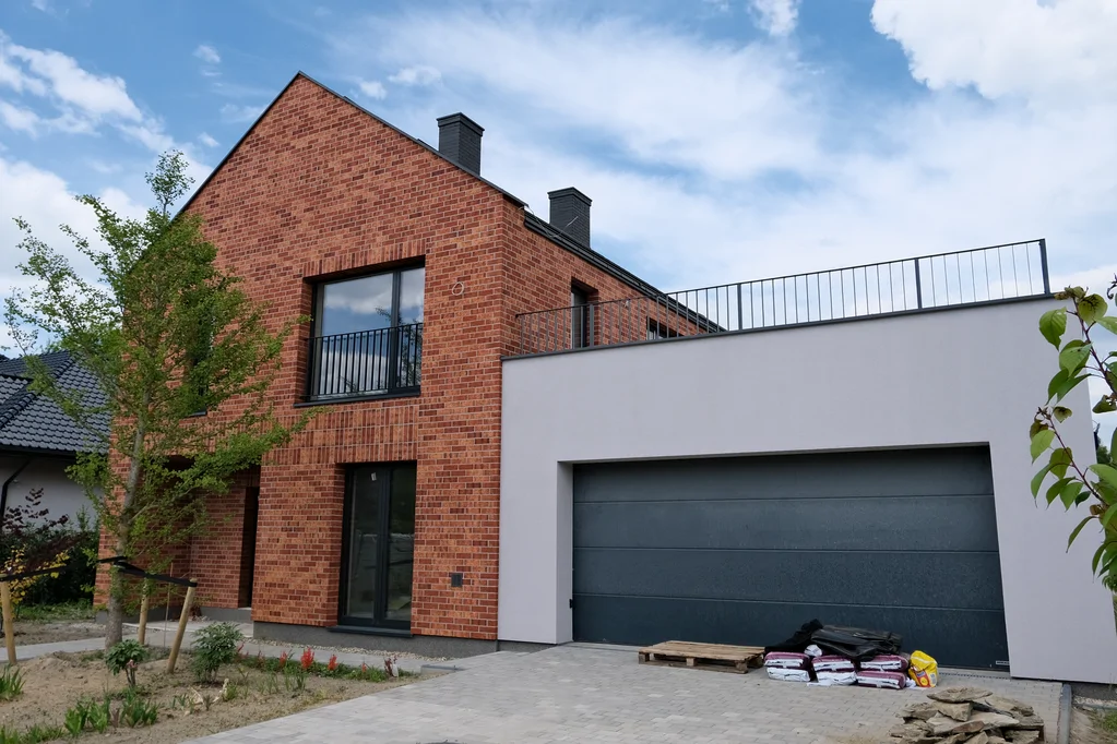 Widok frontowej elewacji domu z ceglaną okładziną, garażem w białej bryle i podjazdem z kostki