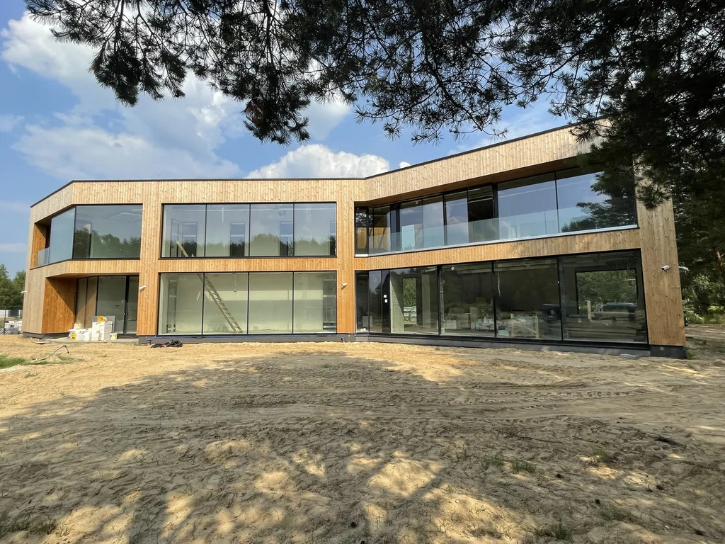 Szeroka elewacja domu z dużymi przeszkleniami na parterze i piętrze; prace końcowe na terenie budowy.