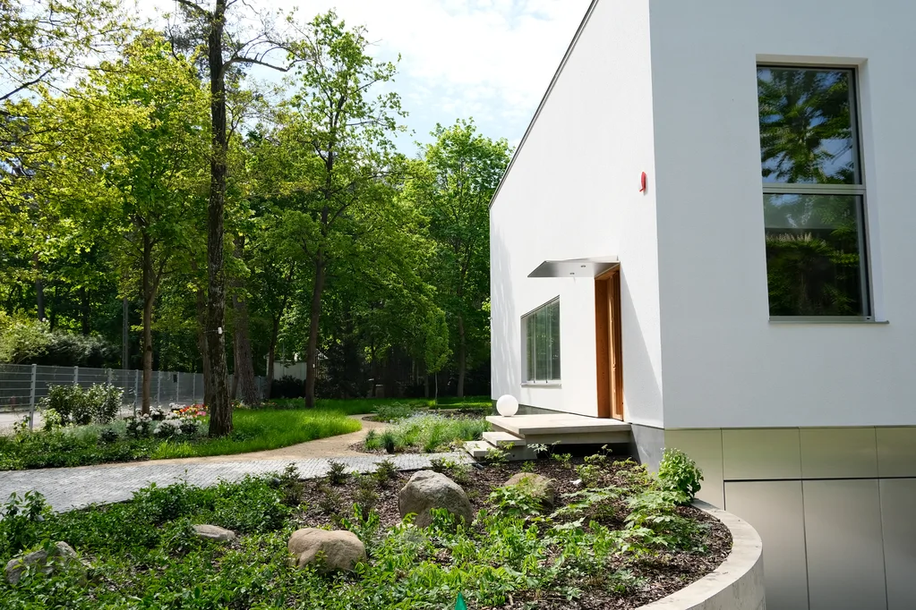 Zbliżenie bocznej elewacji domu z wejściem, ścieżką ogrodową i nasadzeniami przy białej ścianie