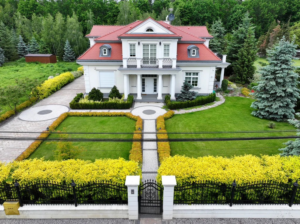 Widok z drona na dom jednorodzinny w stylu dworkowym z białą elewacją z boniami, kolumny okrągłe w strefie wejścia, na balkonie również kolumny i stalowa barierka, pokrycie dachu karpiówka, na dachu widoczne lukarny, przy domu kostka,za domem drzewa,. przed domem widoczne ogrodzenie 