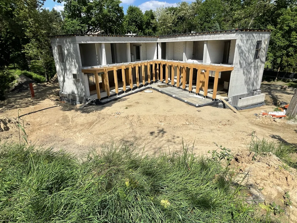 Szeroki widok domu jednorodzinnego w stanie surowym z tarasem i drewnianą pergolą od strony ogrodu