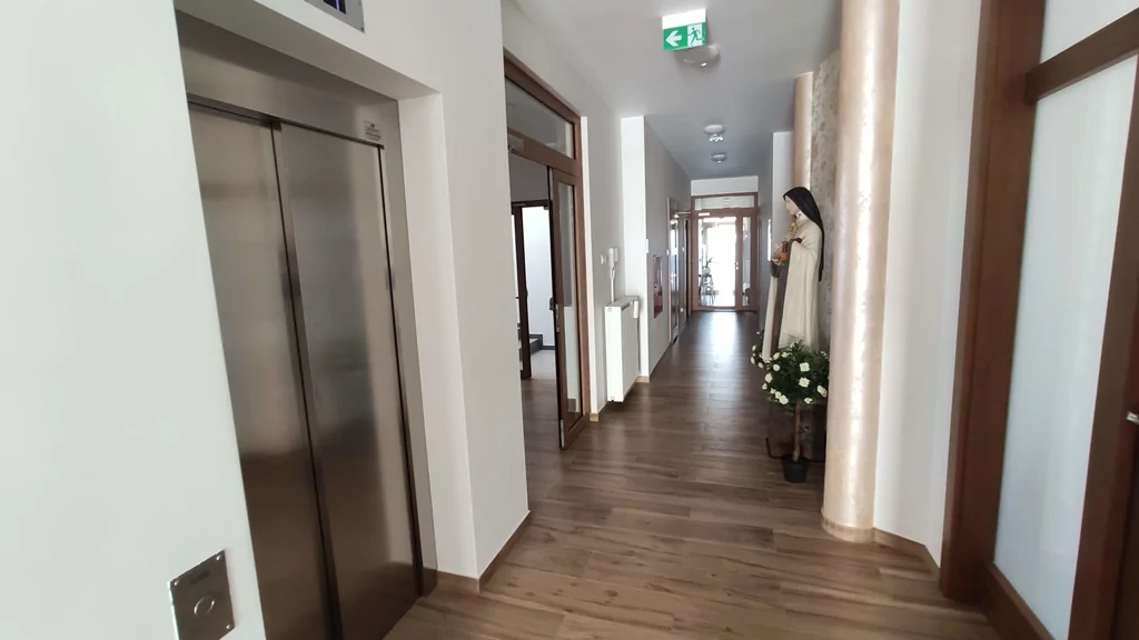 Dom zakonny wewnątrz, widoczna winda, przeszklone drzwi, kolumny ozdobne figurka Maryi 