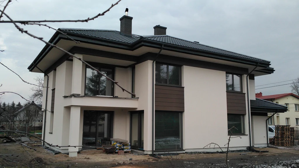 Nowoczesny dom jednorodzinny w trakcie wykończenia, z jasną elewacją, ciemnym dachem kopertowym i dużymi oknami, sfotografowany z ogrodu w pochmurny dzień.