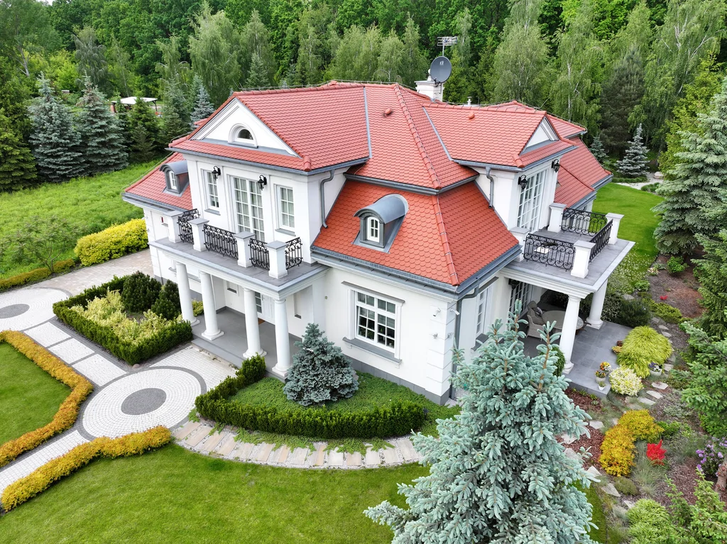 Widok z drona na dom jednorodzinny w stylu dworkowym z białą elewacją z boniami, kolumny okrągłe w strefie wejścia, na balkonie również kolumny i stalowa barierka, pokrycie dachu karpiówka, na dachu widoczne lukarny, przy domu kostka, działka otoczona drzewami 
