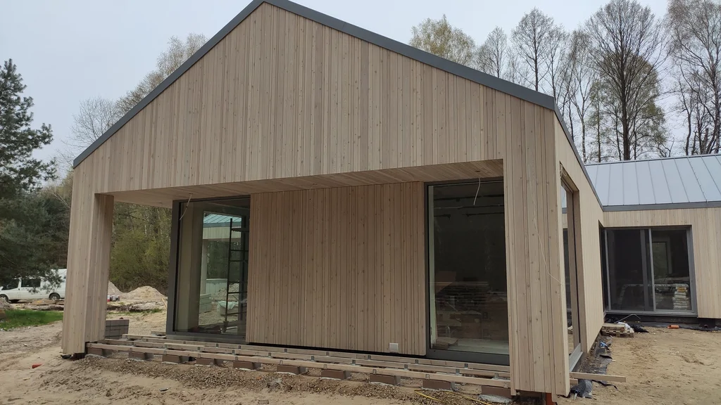 Front domu w trakcie budowy z drewnianą elewacją i dużymi przeszkleniami