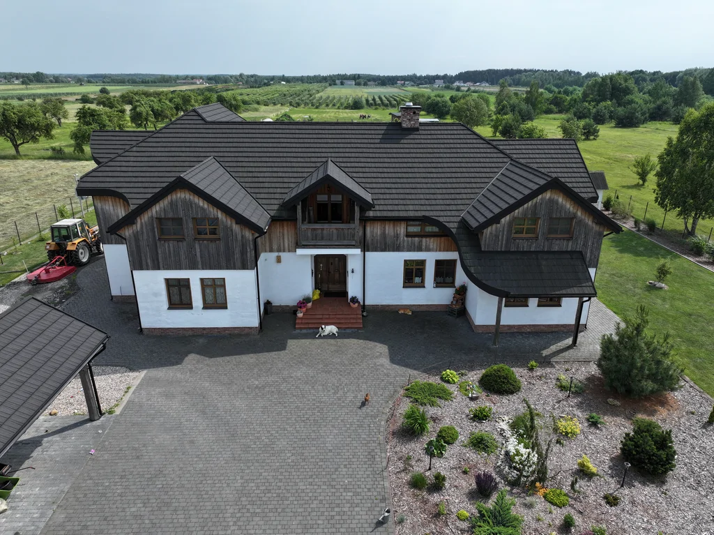 Front domu widziany z drona: centralne wejście i balkon, przed domem podjazd z kostki, w tle pola