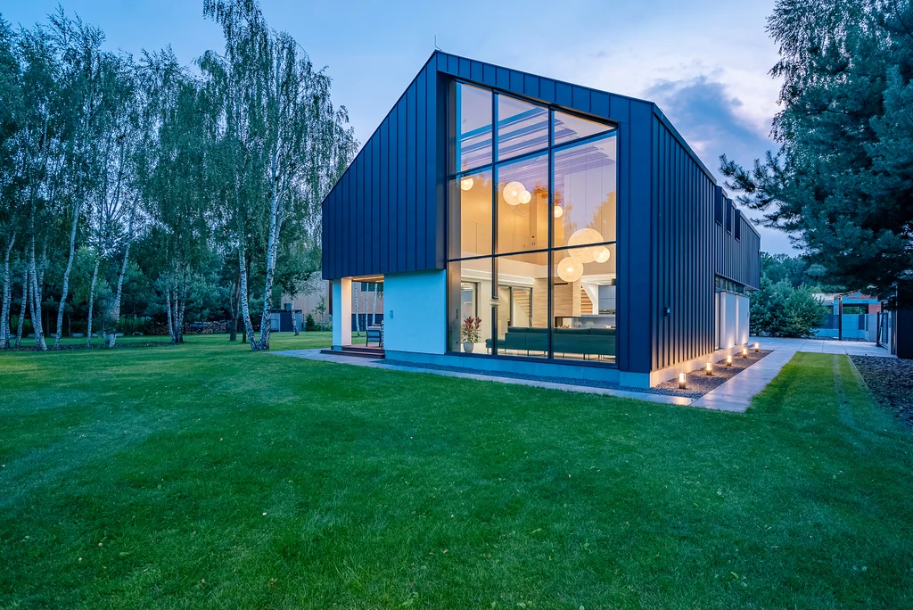 Nowoczesny dom jednorodzinny o zmierzchu, ujęcie narożne od strony ogrodu z wysokim przeszkleniem i podświetloną elewacją.