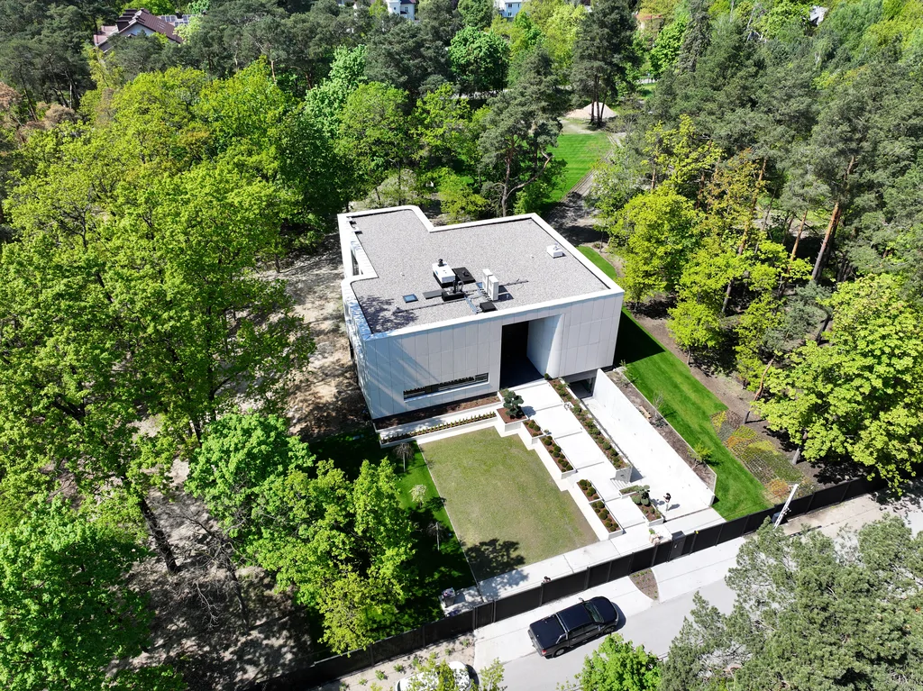 Ukośny widok z drona na bryłę domu, frontowy ogród i otaczające posesję wysokie drzewa