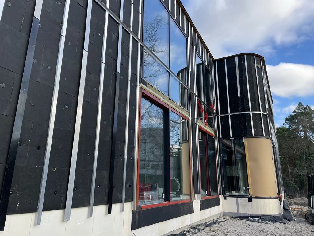 Zaokrąglony narożnik elewacji domu w stanie deweloperskim z dużymi oknami i pionowymi profilami elewacyjnymi