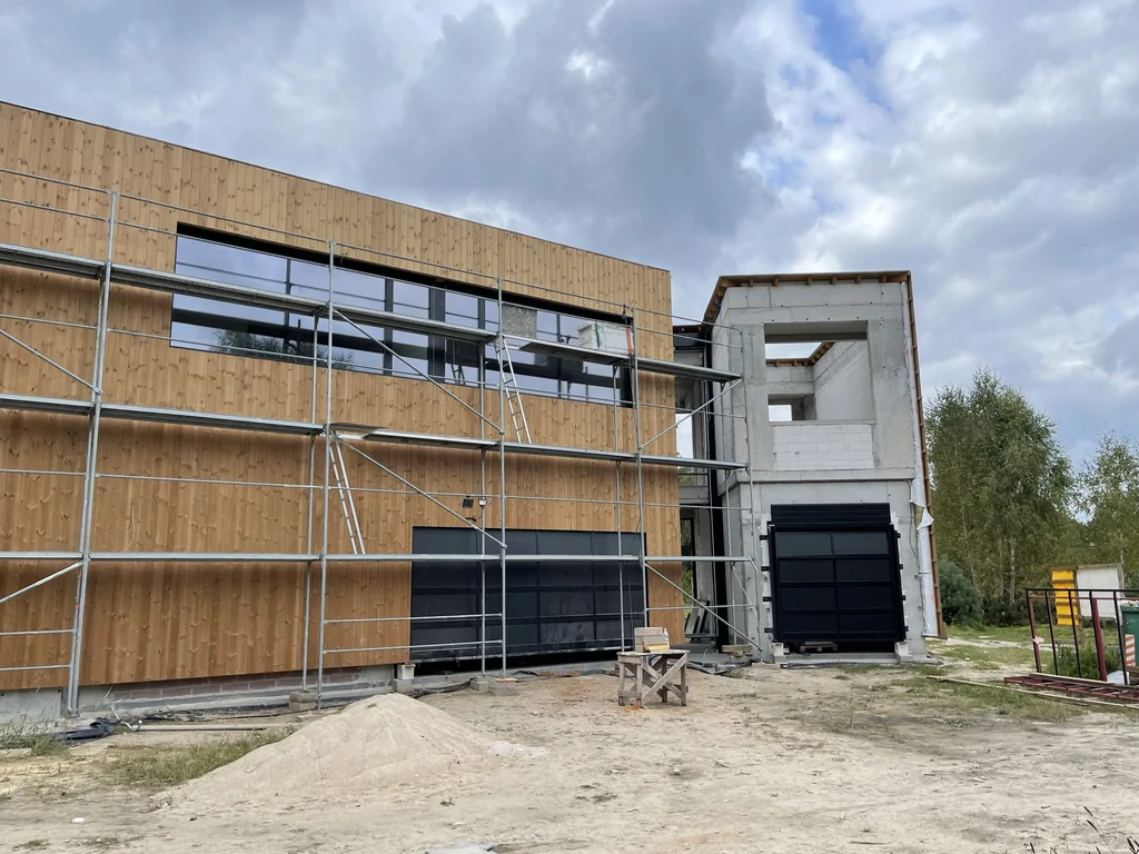 Fragment bryły z widoczną konstrukcją żelbetową i bramą garażową; obok elewacja z pionowego deskowania.